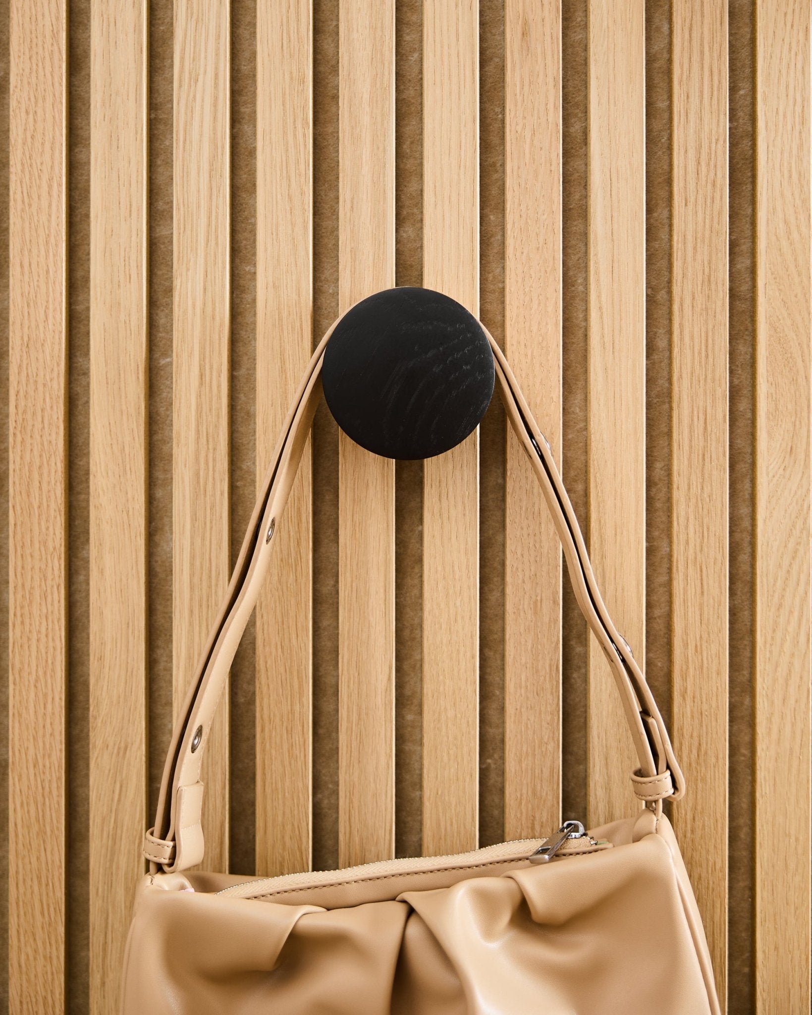 Un bolso Beige colgado de un Linn | Single Hanger de WoodUpp en Black Oak, montado sobre un panel acústico de lamas de madera clara Akupanel.